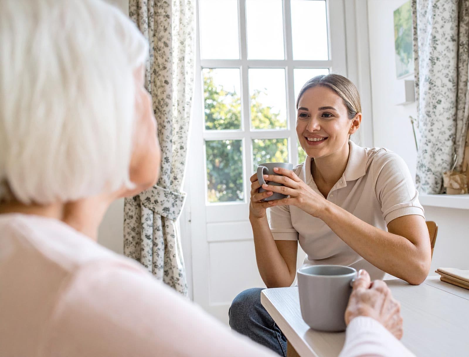 Pflegekraft und Seniorin sitzen am Tisch und trinken aus einer grauen Tassen