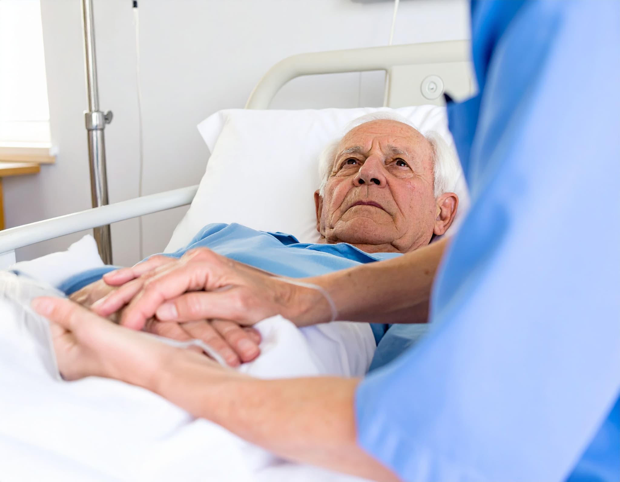 Senior liegt im Krankenhausbett, eine Pflegekraft hält seine Hände fürsorglich