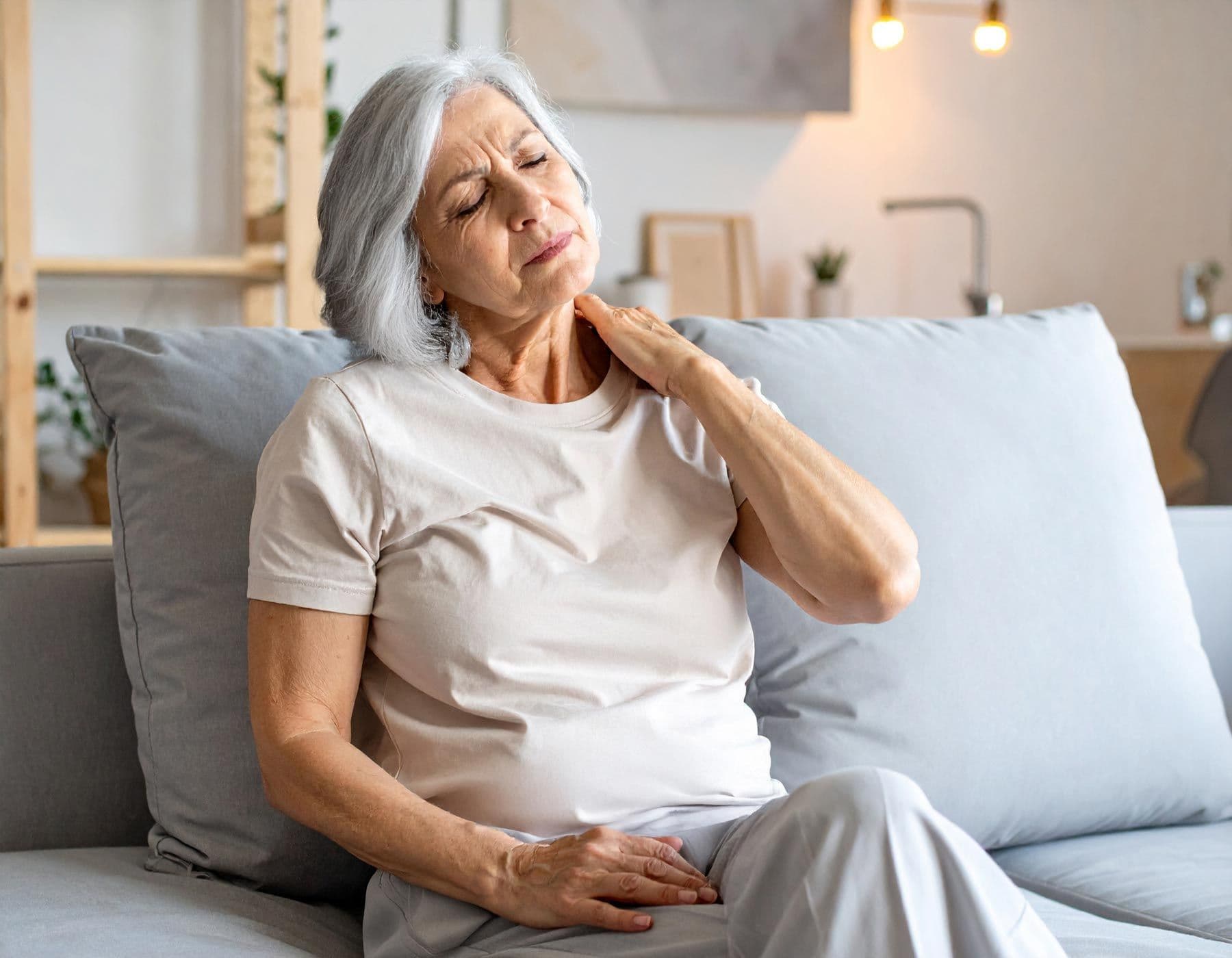 Seniorin sitzt auf Sofa und hält sich schmerzhaft den Nacken