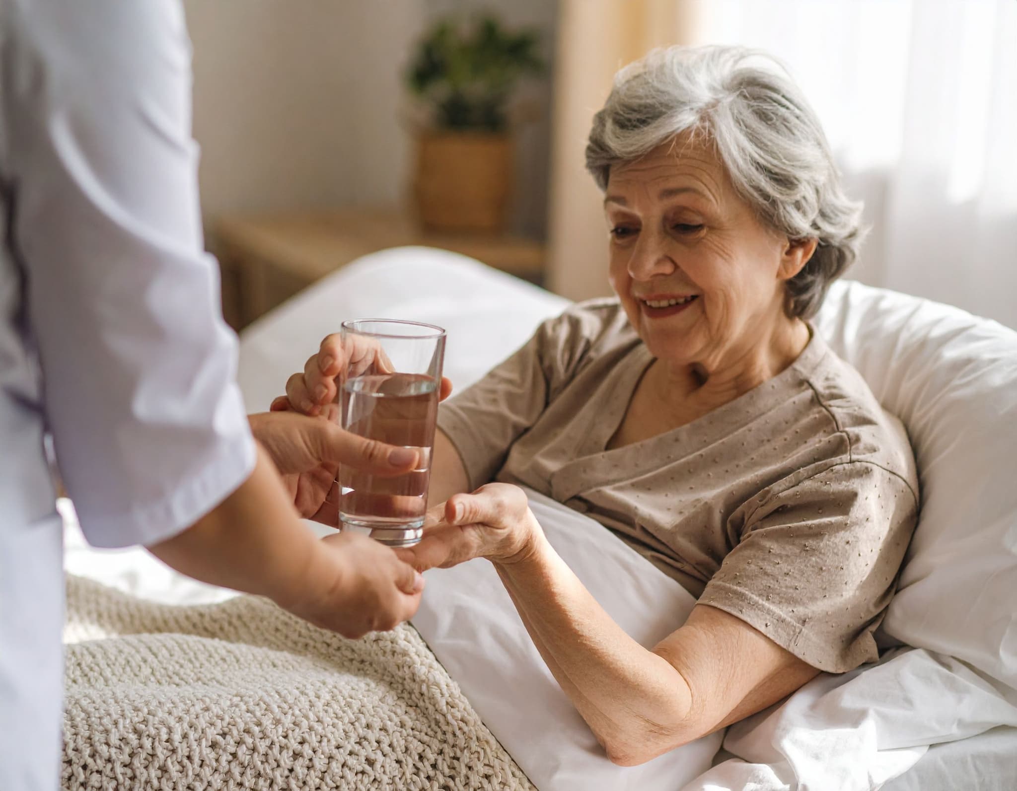 Pflegekraft reicht einer Seniorin im Bett ein Glas Wasser