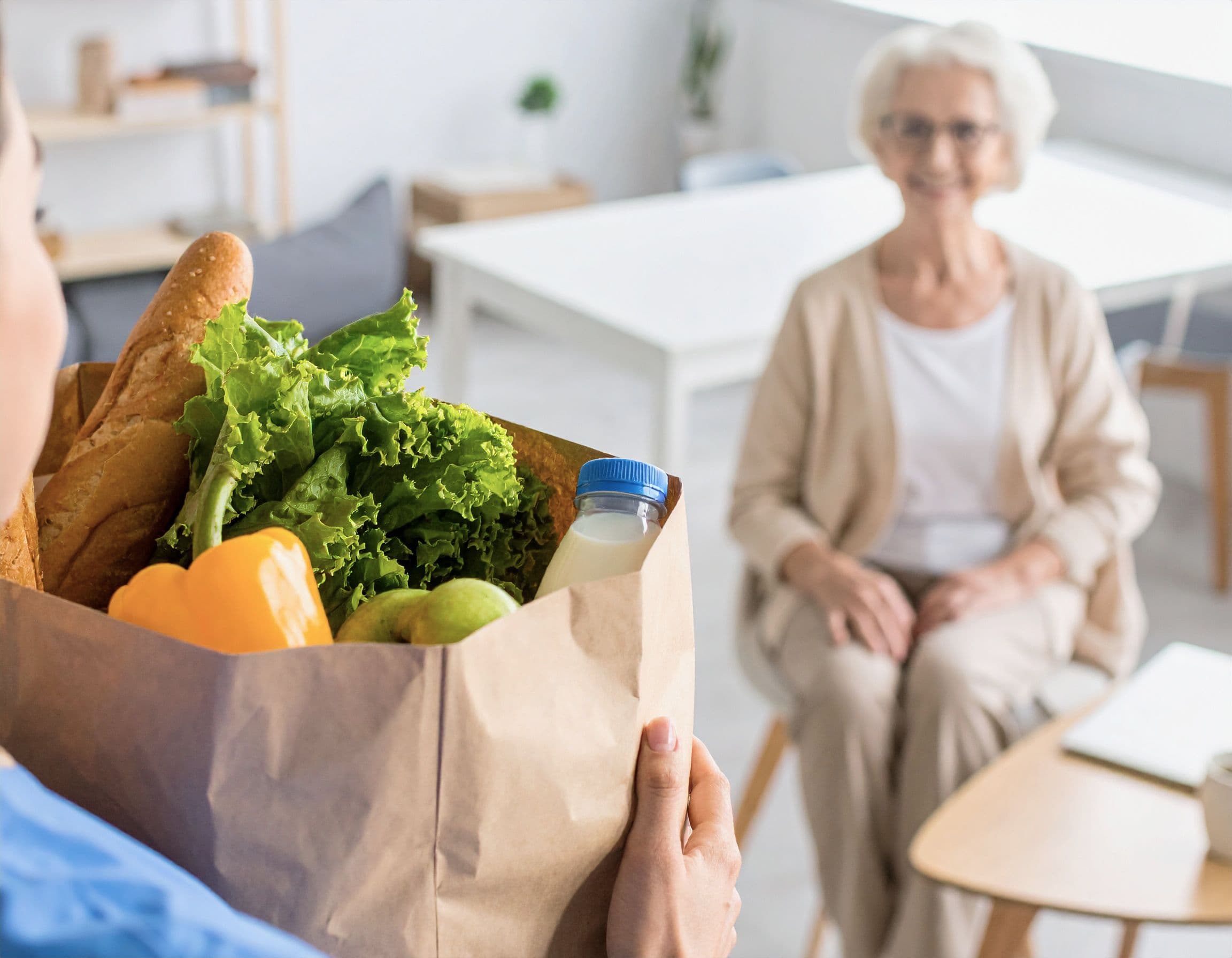 Pflegekraft hält Papiertüte mit frischen Lebensmitteln vor einer Seniorin