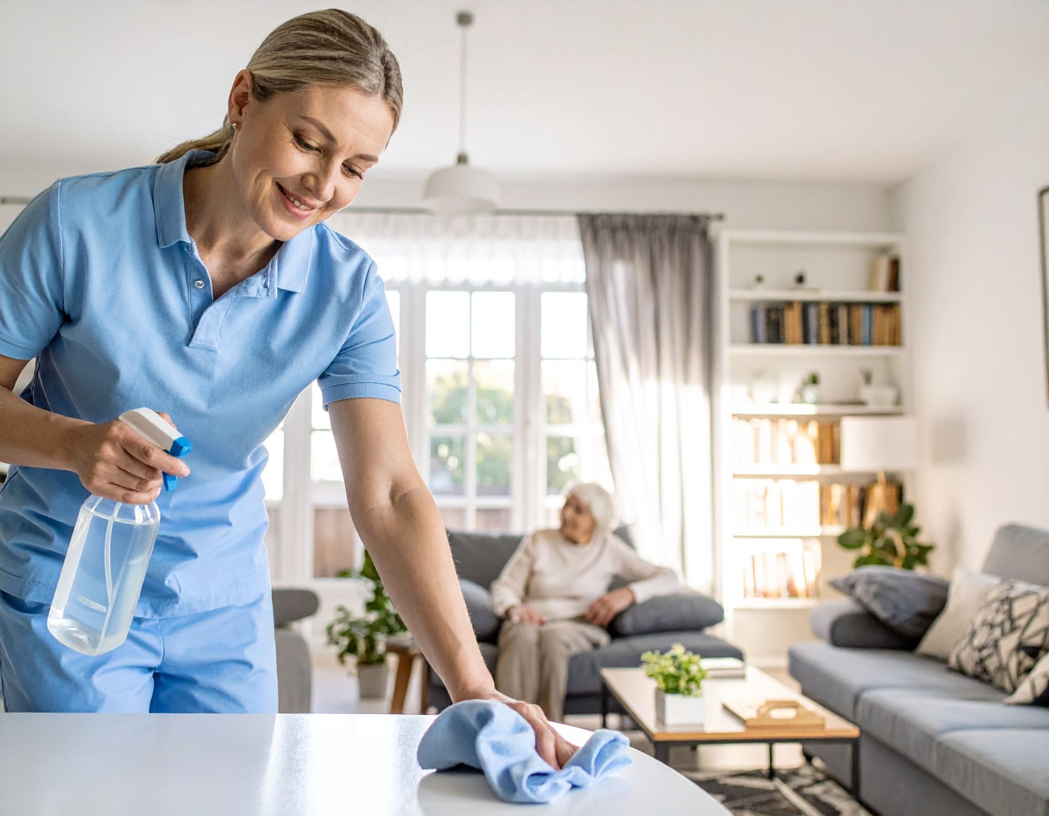 Pflegekraft wischt einen Tisch, ältere Frau sitzt im Hintergrund auf einem Sessel