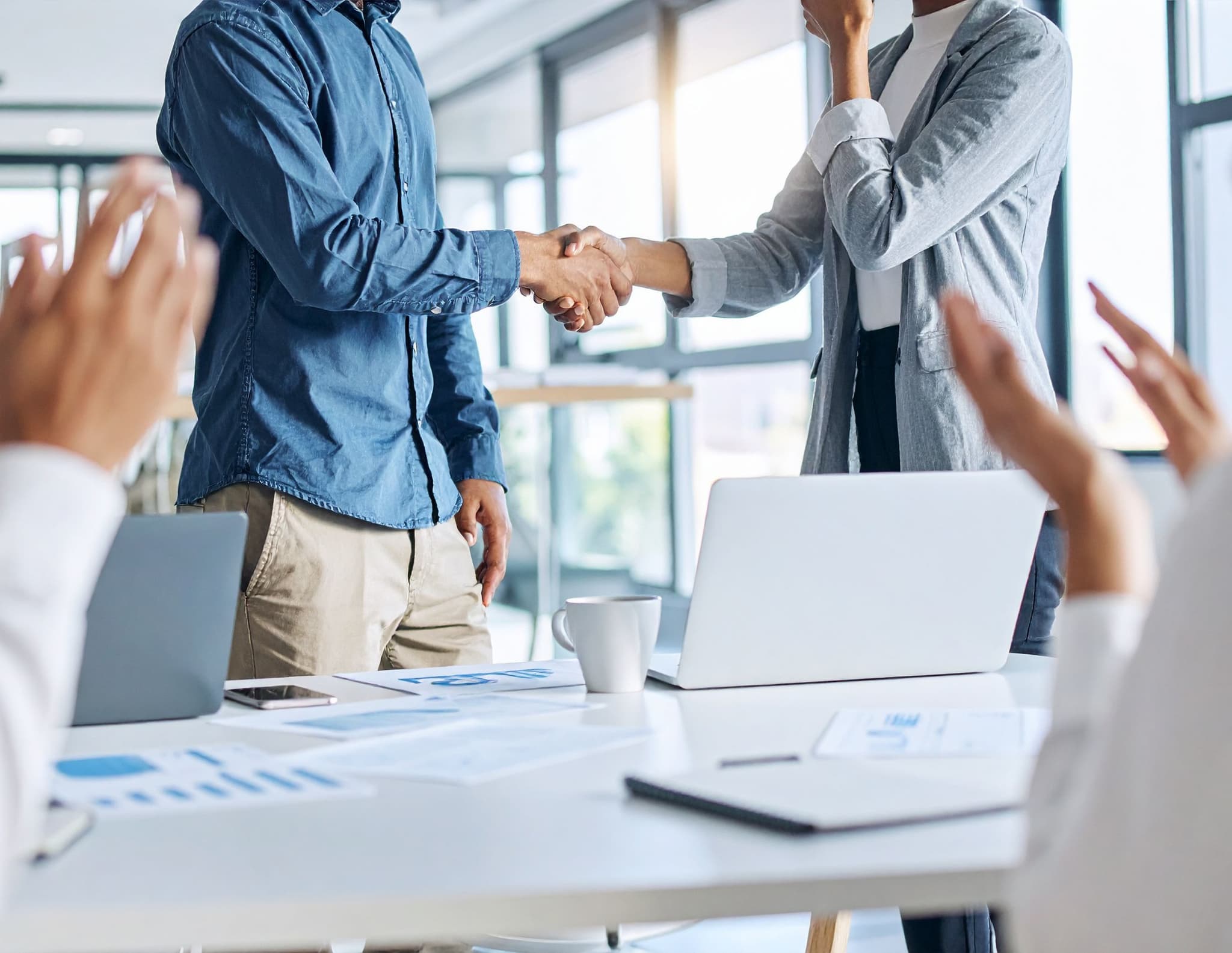 Zwei Personen stehen in einem Büro und schütteln sich die Hand, während andere applaudieren