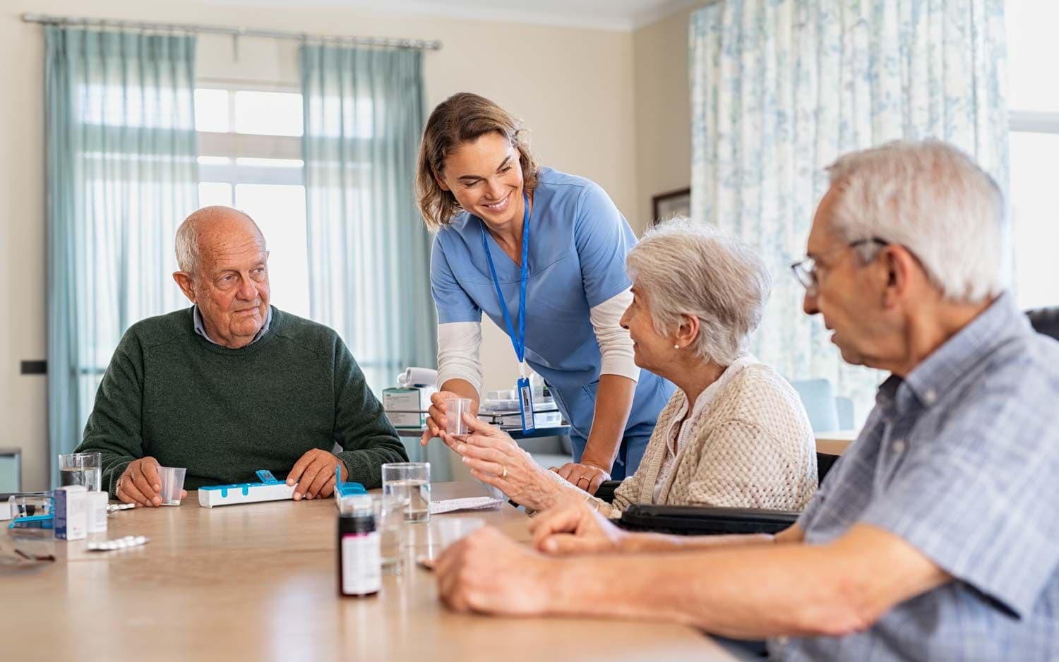 Pflegekraft reicht älterer Frau am Tisch ein Medikament, zwei ältere Männer sitzen daneben