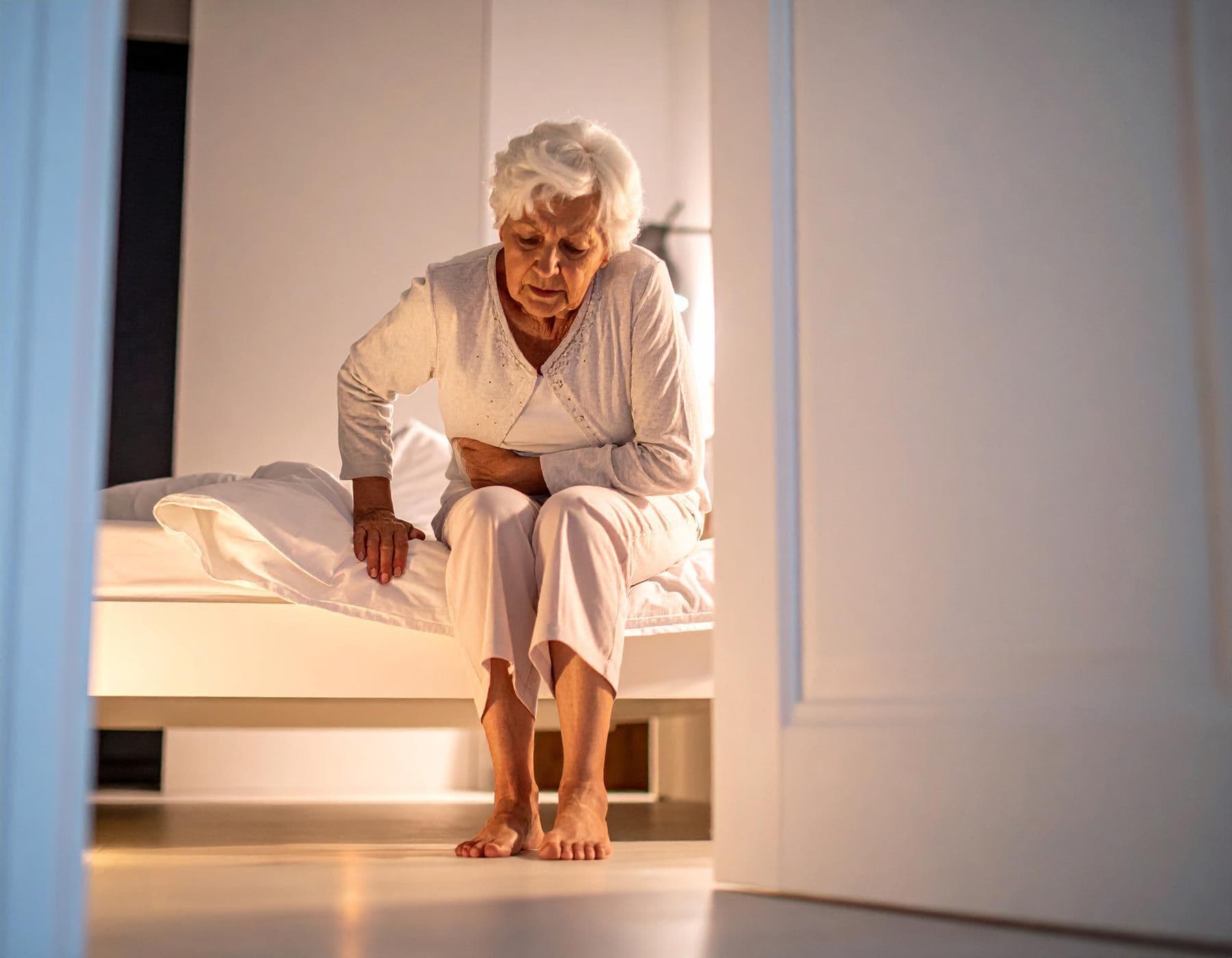 Ältere Frau sitzt auf Bettkante und hält schmerzhaft ihren Bauch