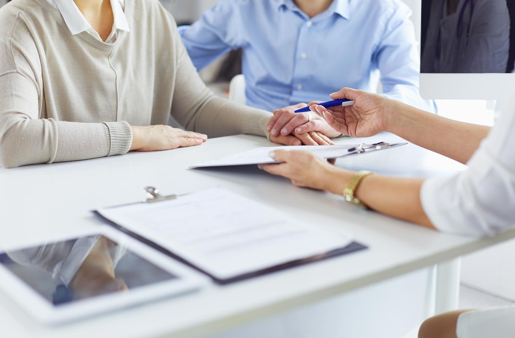 Zwei Personen sitzen an einem Tisch, eine dritte Person reicht einen Stift und hält ein Klemmbrett mit Dokumenten