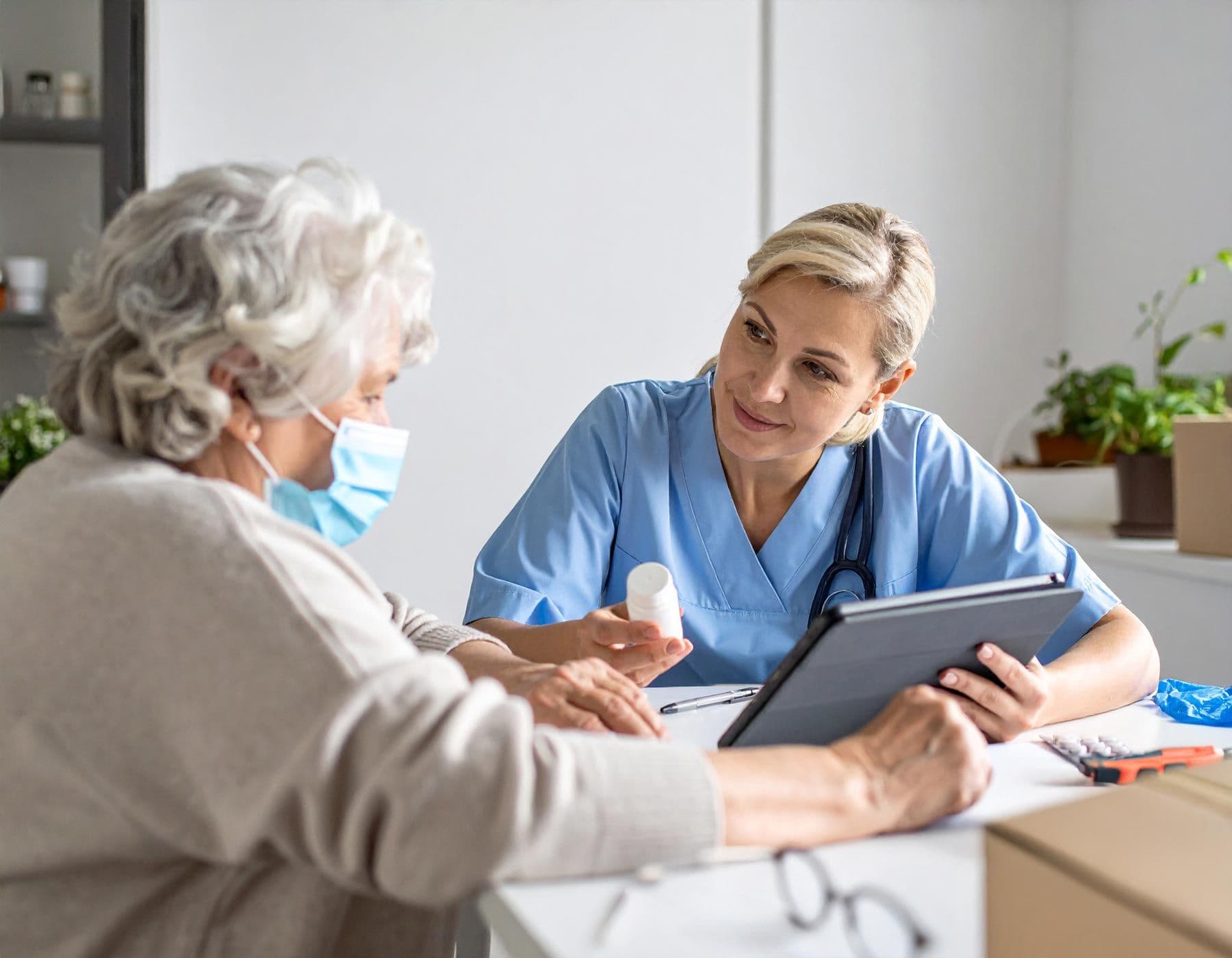 Pflegekraft sitzt am Tisch mit älterer Frau und hält ein Tablet, zeigt ihr eine Medikamentenflasche