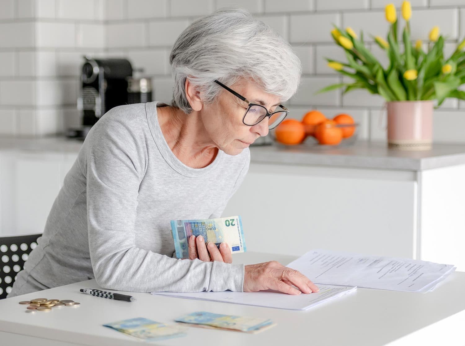 Ältere Frau sitzt am Tisch, hält Euro-Geldscheine und betrachtet Dokumente in moderner Küche