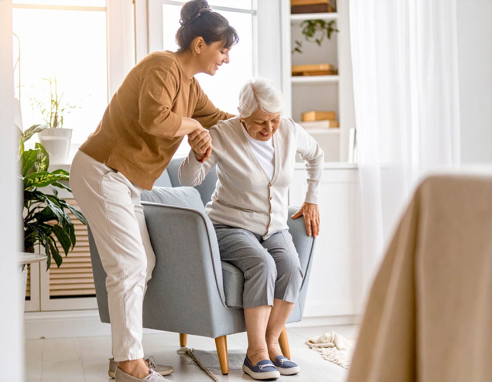 Seniorin sitzt im Sessel, wird von einer anderen erwachsenen Frau beim Aufstehen unterstützt