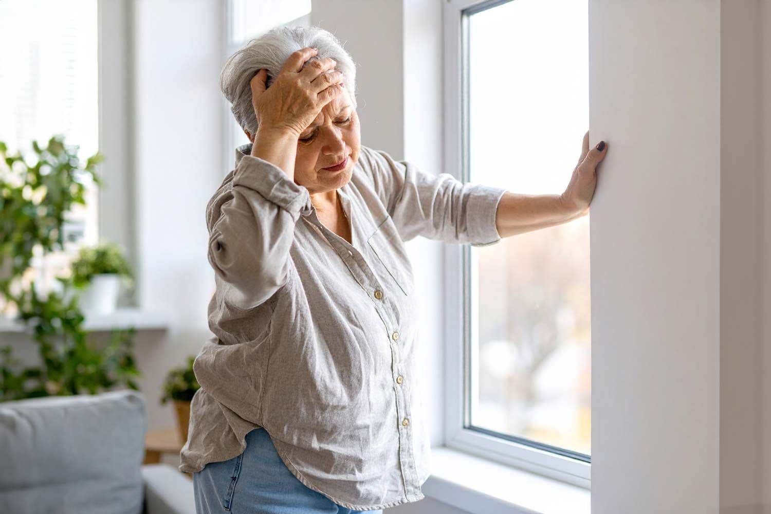 Ältere Frau lehnt sich an eine Wand neben einem Fenster, hält sich den Kopf