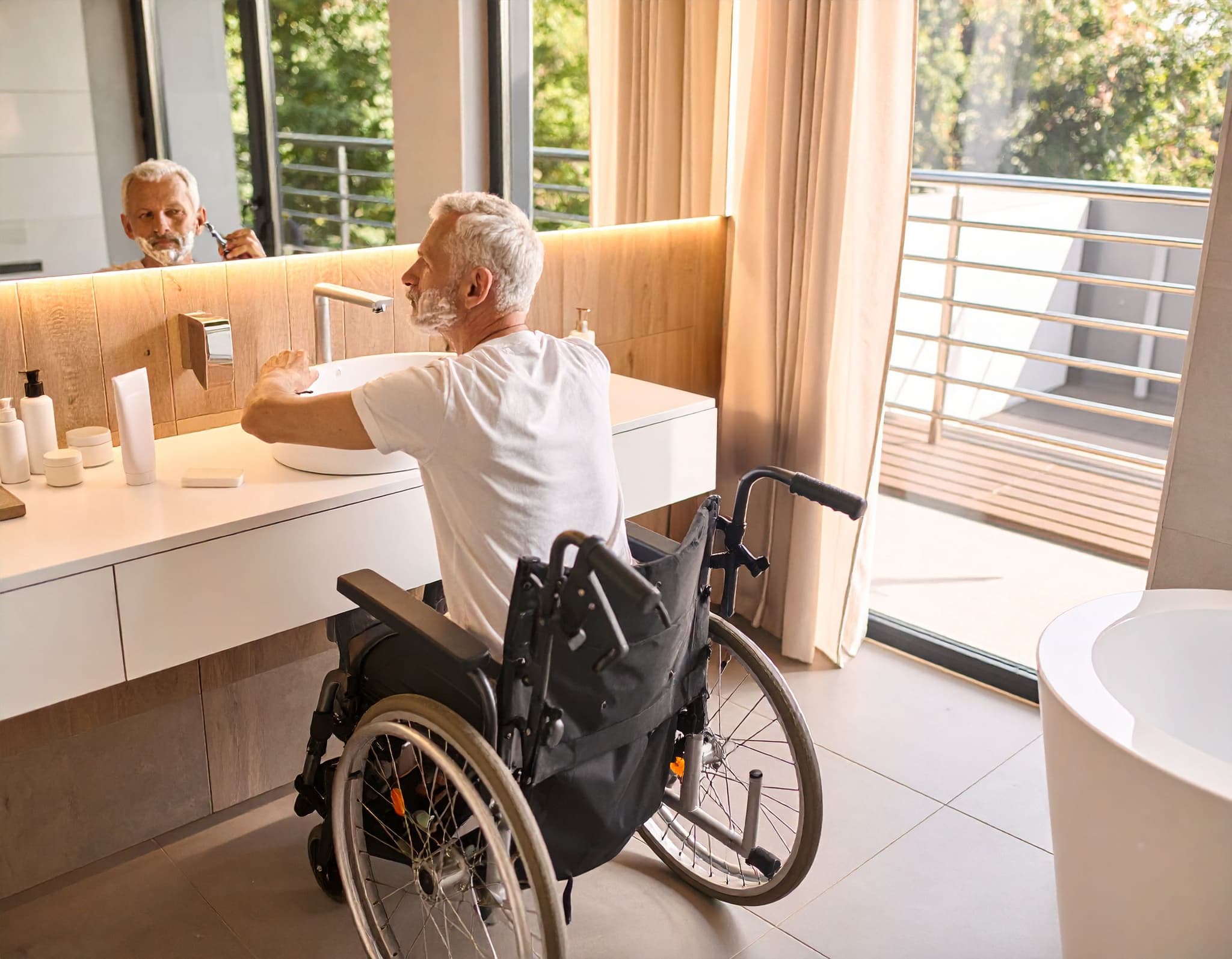 Senior im Rollstuhl sitzt vor Waschbecken und rasiert sich im hellen Badezimmer