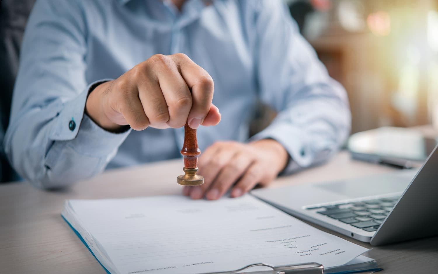 Person stempelt ein Dokument am Schreibtisch neben einem Laptop
